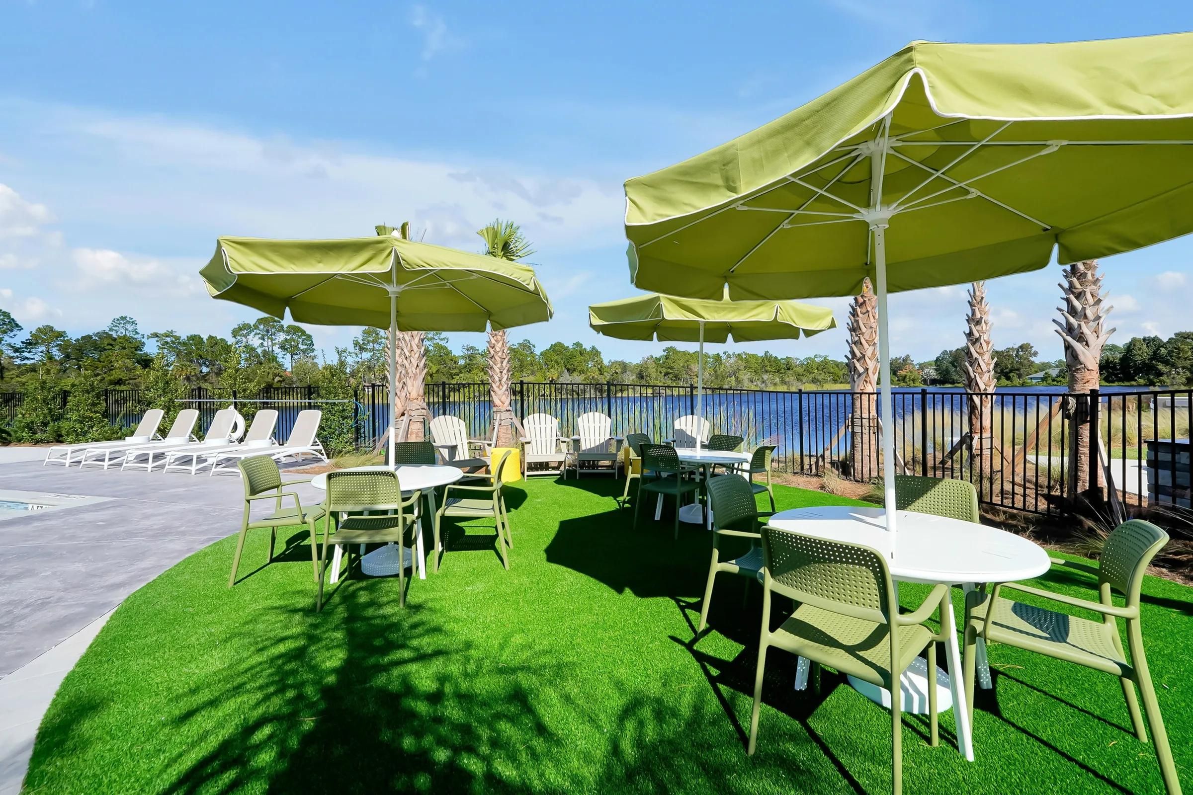 Outdoor seating area with green umbrellas, tables, and chairs, overlooking water and palm trees on a sunny day.