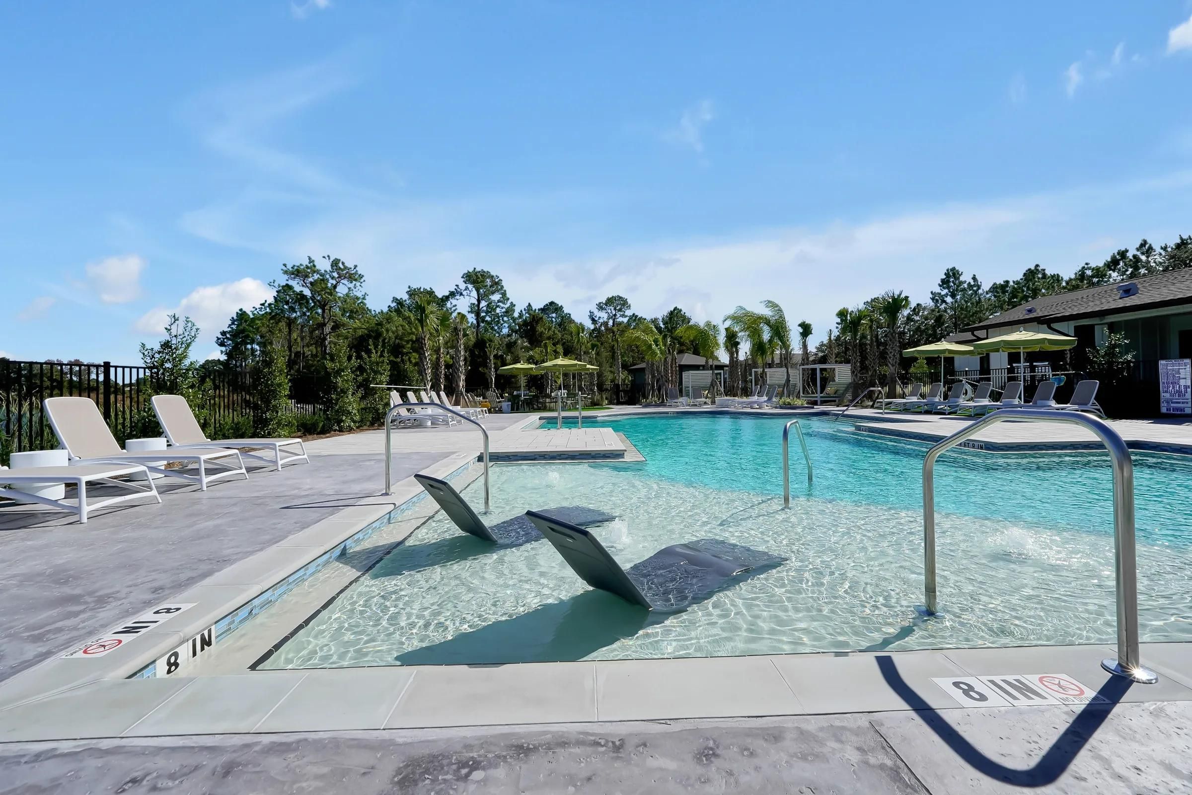 Outdoor swimming pool with lounge chairs in the shallow end, surrounded by trees and umbrellas.