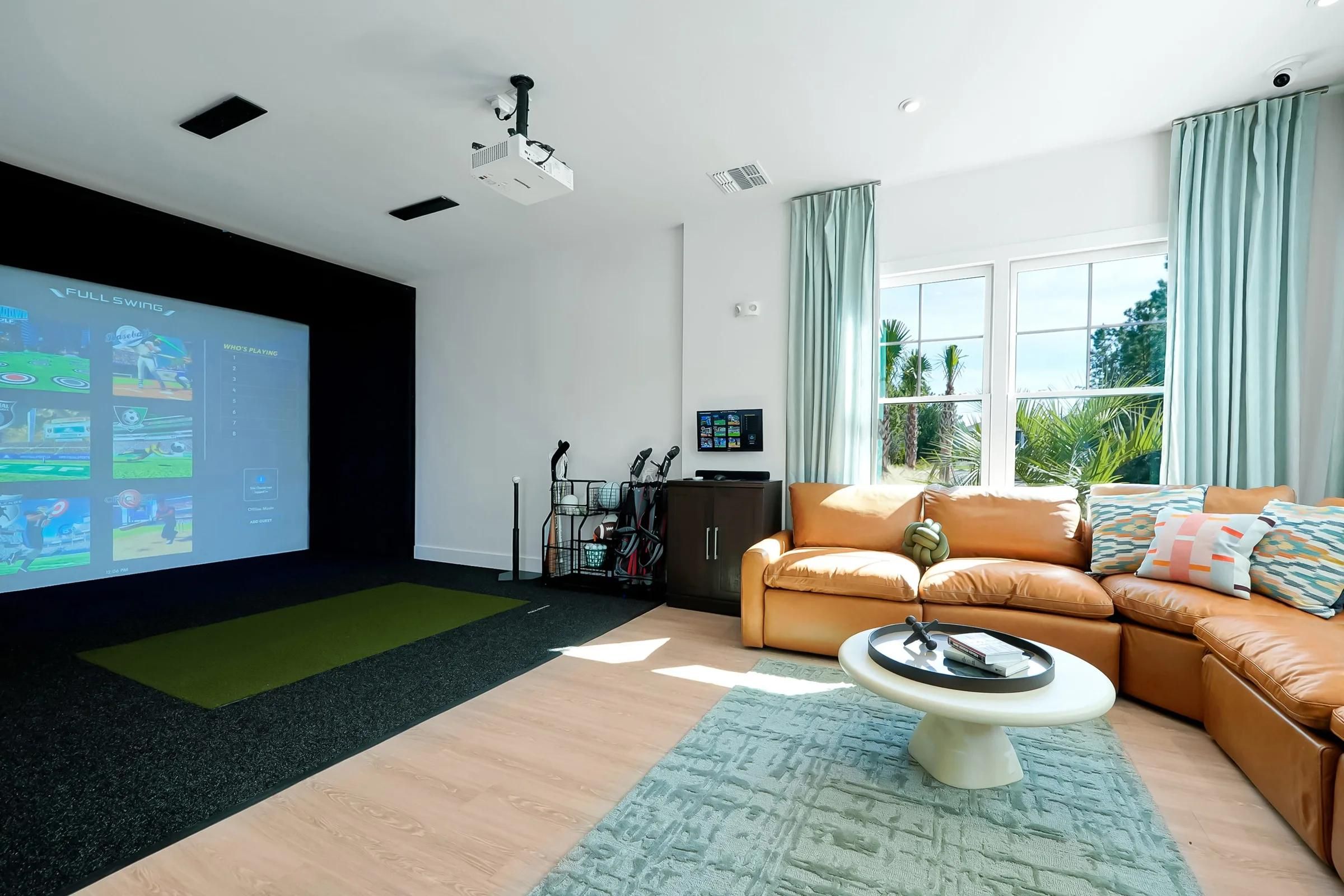 Modern room with a golf simulator, tan sofa, round table, clubs in corner, and large windows.