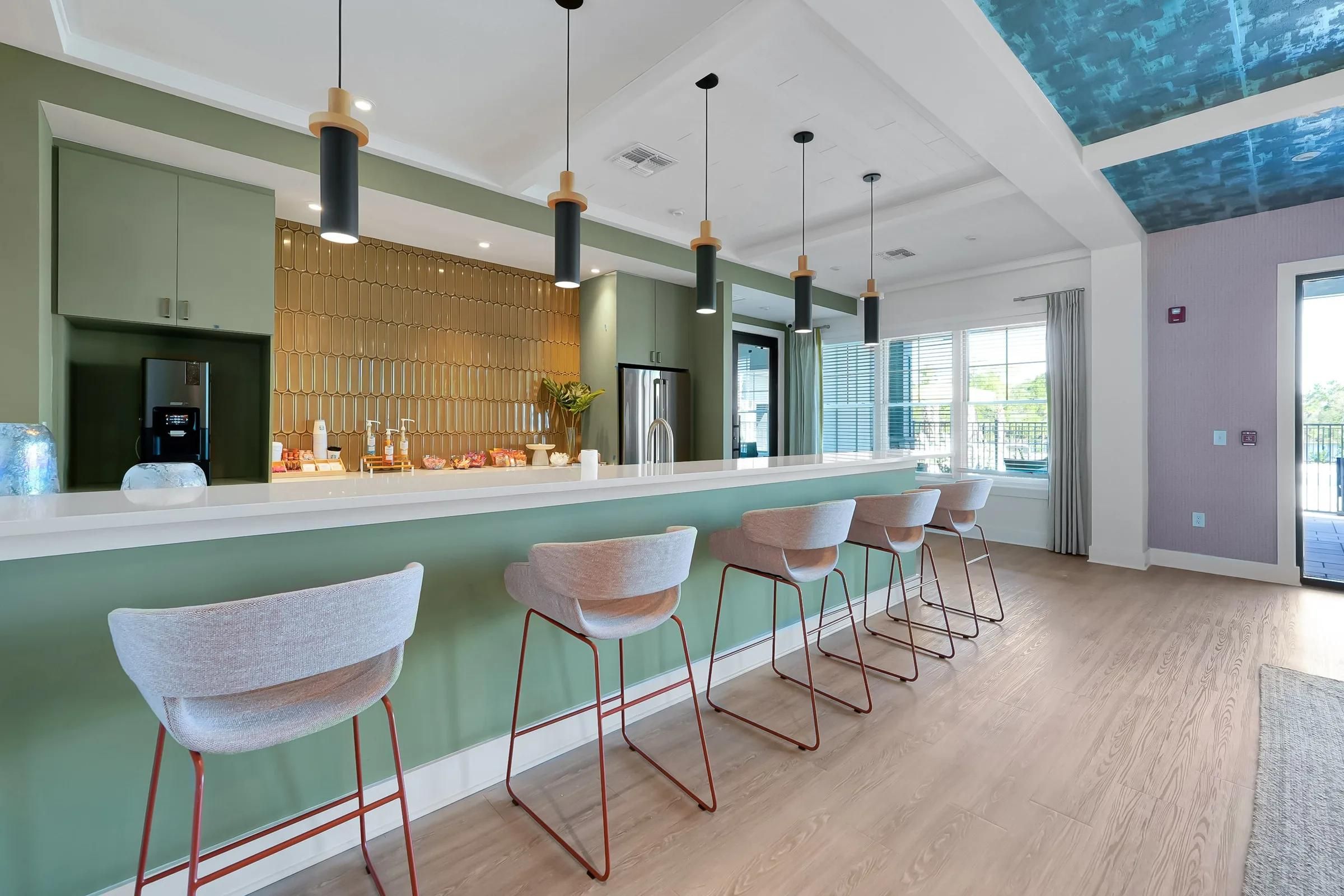 Modern bar with green accents, gold backsplash, hanging lights, and five bar stools in a bright, airy space.